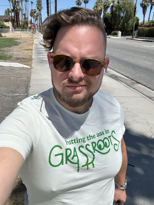 Selfie outdoors in Palm Springs wearing sunglasses and a ‘Putting the Ass in Grassroots’ t-shirt, palm trees in background